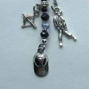 May include: A silver-toned charm accessory featuring a central strand of black and light purple faceted beads. Attached are three distinct silver-colored metal charms: a baseball cap, a crossbow, and a standing figure in a long-sleeved shirt.
