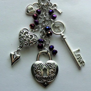 May include: A silver-toned charm cluster featuring heart and key pendants. The charms include a heart-shaped padlock, a key with the word "love", and a key with a star cut-out. Dark purple beads are interspersed throughout the chain.