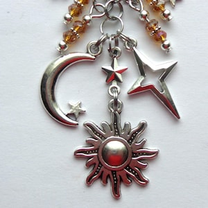 May include: A celestial-themed charm pendant featuring silver-toned charms of a crescent moon, stars, and a sunburst. The pendant is accented with amber-colored beads and is designed to be attached to a necklace or bracelet.