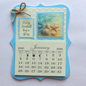 May include: A handmade calendar for January 2026, featuring a beach theme. The calendar has a light blue frame with a decorative edge. It includes a picture of seashells and the text "Every Seashell has a story."