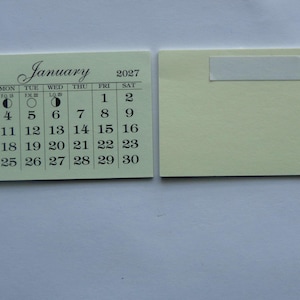 May include: A small, pale green calendar card for January 2027, with the month's name in elegant script. The calendar grid features dates, days of the week, and moon phases. A second card with adhesive is next to it.