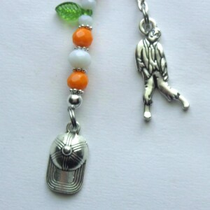May include: A metal bookmark featuring a vertical strand of alternating orange and white beads, topped with a small green leaf charm. Attached to the bottom are two silver-toned metal charms: one shaped like a baseball cap and one shaped like a person.