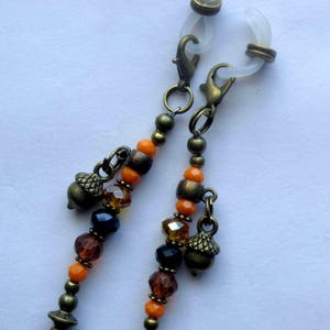 May include: A pair of body jewelry rings featuring bronze-toned metal accents, orange and dark brown faceted beads, and small bronze acorn charms. The jewelry is attached to clear, flexible silicone rings with bronze-toned lobster claw clasps.