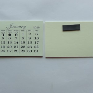 May include: A white calendar with a black magnet on the back. The calendar is for January 2026 and shows the days of the week and the dates. The magnet is a rectangular shape and is attached to the back of the calendar.