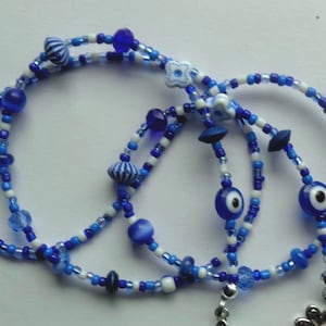 May include: A beaded necklace featuring a mix of blue and white beads. The necklace includes various bead shapes, including round, striped, and eye-shaped beads. A silver-colored charm hangs at the bottom. The necklace is designed with multiple strands.