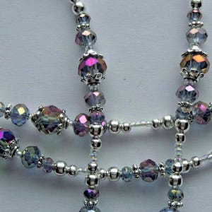 May include: Close-up of a handmade necklace featuring iridescent crystal beads in shades of purple, blue, and gold, interspersed with small silver beads. The necklace has a delicate, intricate design.