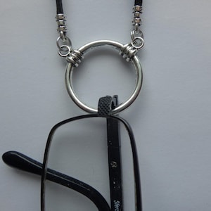 May include: A silver-colored eyeglass holder with a black cord. The holder features a large circular ring and is designed to hold eyeglasses. The eyeglasses have black frames.