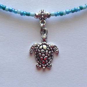 May include: A silver turtle charm hangs from a blue and white beaded necklace. The turtle charm has a detailed shell and is a popular symbol of good luck and protection.