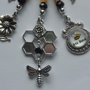 May include: A collection of silver-toned bee-themed charms. The charms include a flower, a honeycomb, and a circular charm with a bee and the words "bee kind." The charms are connected with silver-toned beads and are likely used for jewelry or crafts.