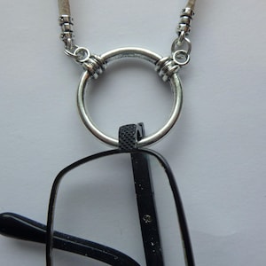 May include: A silver-toned metal ring pendant attached to a chain, designed to hold a pair of black eyeglasses. The circular ring features decorative ribbed detailing at the top where it connects to the chain, and the black glasses hang vertically.