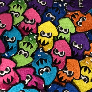 Könnte beinhalten: Nahaufnahme einer Sammlung gestickter Tintenfisch-Patches in einer Vielzahl von leuchtenden Farben, darunter Rosa, Blau, Grün, Gelb, Orange und Lila. Jeder Patch hat eine schwarze Umrandung und ein weißes Auge. Die Patches sind dicht gepackt.
