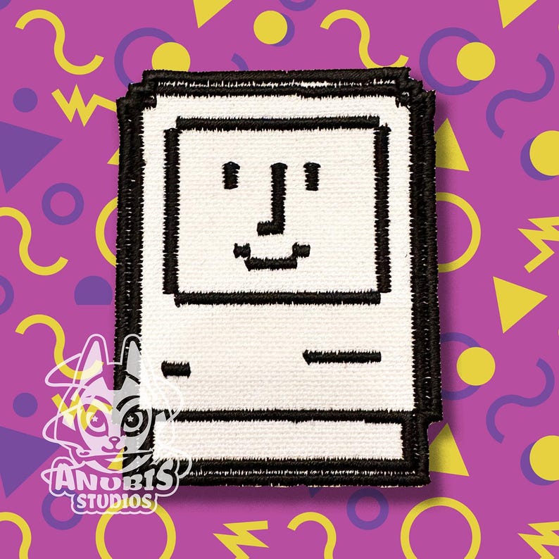 May include: A white embroidered patch depicting a vintage computer with a smiling face. The patch has a black outline and is surrounded by a black border.