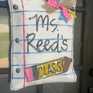 May include: A handmade door sign resembling a lined notebook page. The sign reads "Ms. Reed's CLASS" with a pencil illustration. A multi-fabric bow with pink polka dots and burlap accents adorns the top. The sign is designed to hang on a door.