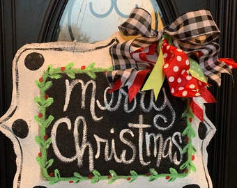 May include: A rectangular Christmas sign with a black background and white lettering that reads "Merry Christmas." The sign is framed in white with black polka dots and a green and red holly border. A large bow with black and white checkered ribbon and red polka dot ribbon is attached.