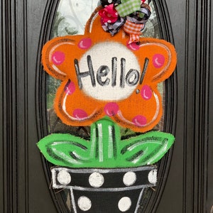 May include: A flower-shaped door hanger with the word "Hello!" in the center. The orange flower has pink polka dots and a green stem and leaves. The flower pot is black with white polka dots, and a colorful bow is at the top.
