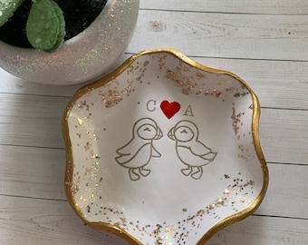 May include: White ceramic trinket dish with a gold rim and gold glitter accents. The dish features two bird illustrations, a red heart, and the letters "C" and "A". A decorative succulent plant in a white pot is in the background.