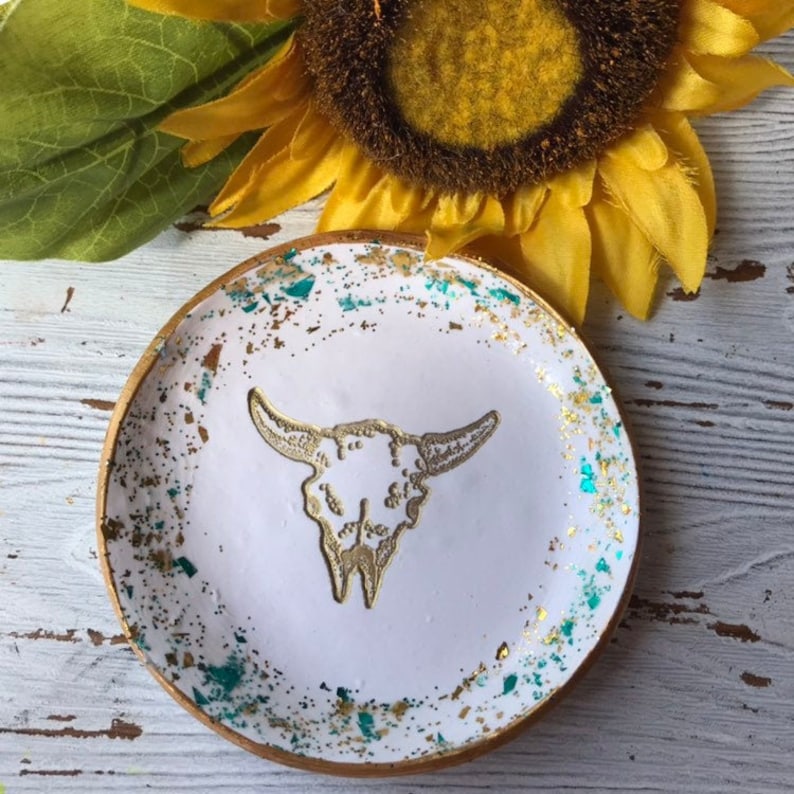 May include: White ceramic ring dish with a gold bull skull design and teal and gold glitter accents.