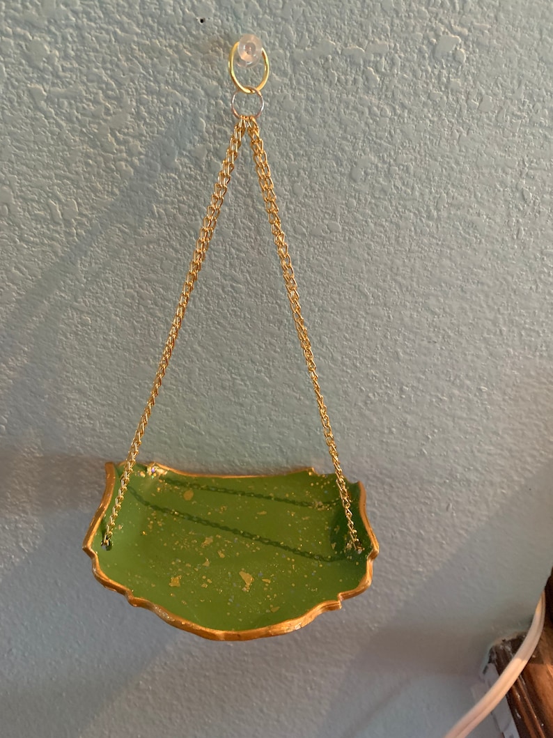 K&ouml;nnte beinhalten: Ein gr&uuml;nes H&auml;ngeregal mit goldenen Verzierungen und einer goldenen Kette. Das Regal h&auml;ngt an einem Haken an einer hellblauen Wand.