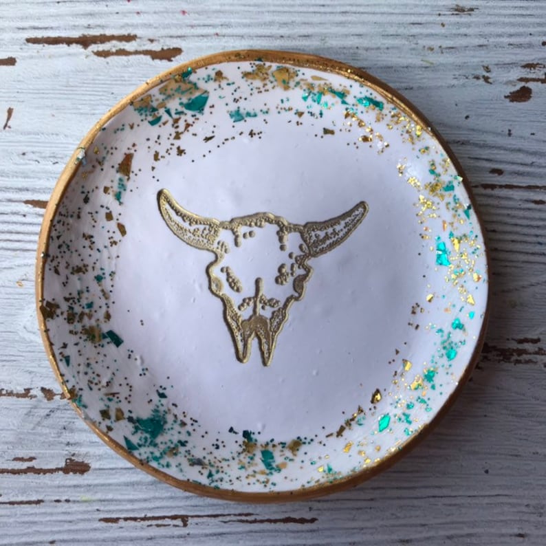 May include: White ceramic trinket dish with a gold rim and a gold glitter design. The dish features a raised gold bull skull design in the center.