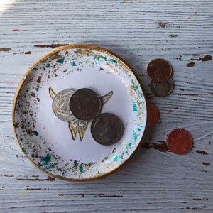 May include: White ceramic trinket dish with a gold rim and a gold and teal glitter design. The dish features a white silhouette of a cow skull with horns. There are five US coins scattered around the dish.
