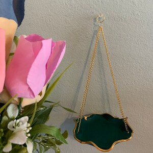 Op de afbeelding: Een groene en gouden hangende plantenpot met een ketting. De pot heeft een golvende rand en hangt aan een gouden ketting.