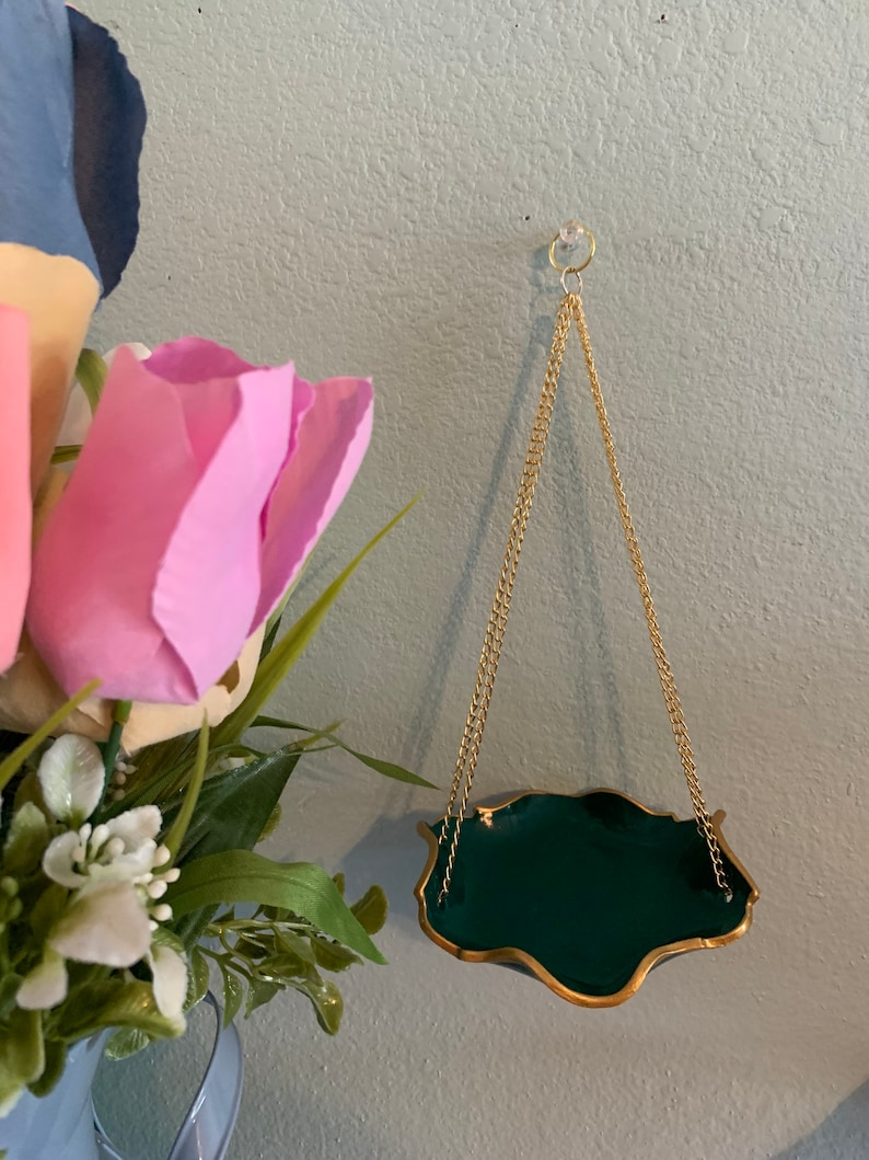 Op de afbeelding: Een groene en gouden hangende plantenpot met een ketting. De pot heeft een golvende rand en hangt aan een gouden ketting.