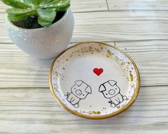 May include: White ceramic trinket dish with a gold rim and gold flecks. The dish has two black and white pig illustrations and a red heart in the center.