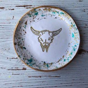 May include: White ceramic trinket dish with a gold rim and a gold glitter design. The dish features a white silhouette of a bull skull.