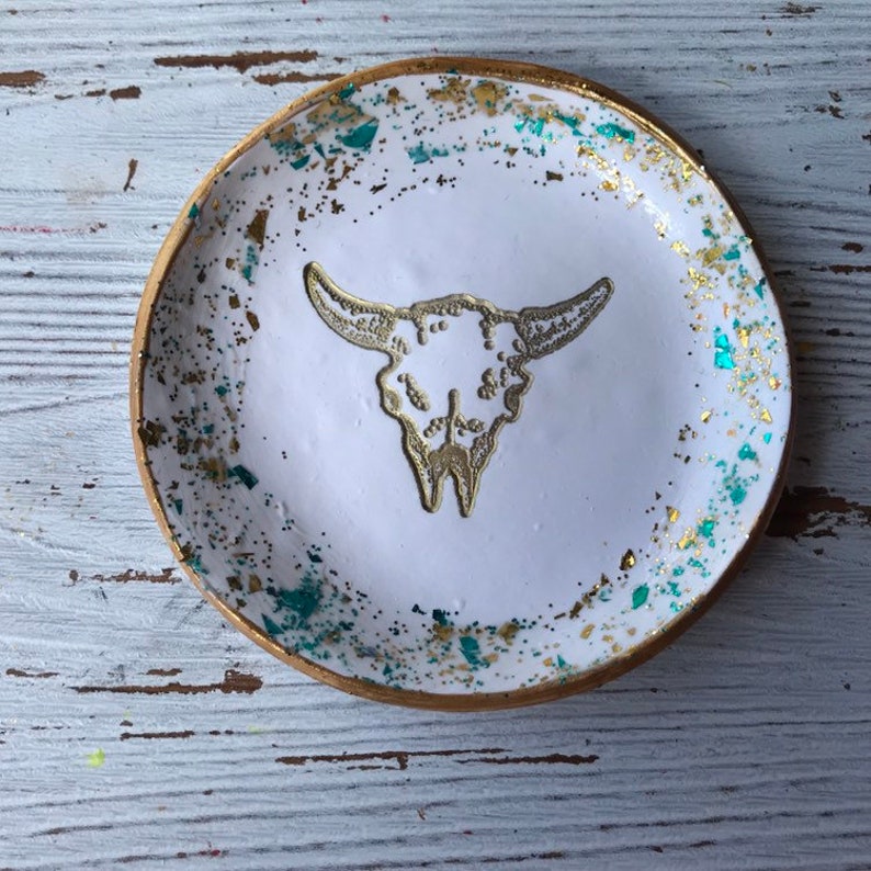May include: White ceramic trinket dish with a gold rim and a gold glitter design. The dish features a white silhouette of a bull skull.