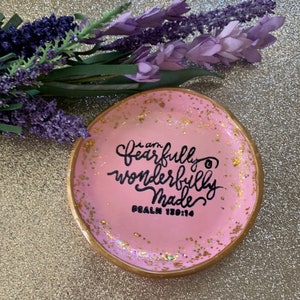 May include: A pink trinket dish with gold glitter and a gold rim. The dish has the words "I am fearfully and wonderfully made" written in black cursive lettering. The words "Psalm 139:14" are written in black below the cursive lettering.