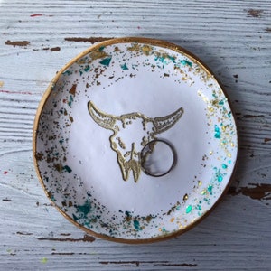 May include: White ceramic ring dish with a gold rim and a gold and teal glitter design. The dish features a white silhouette of a bull skull.