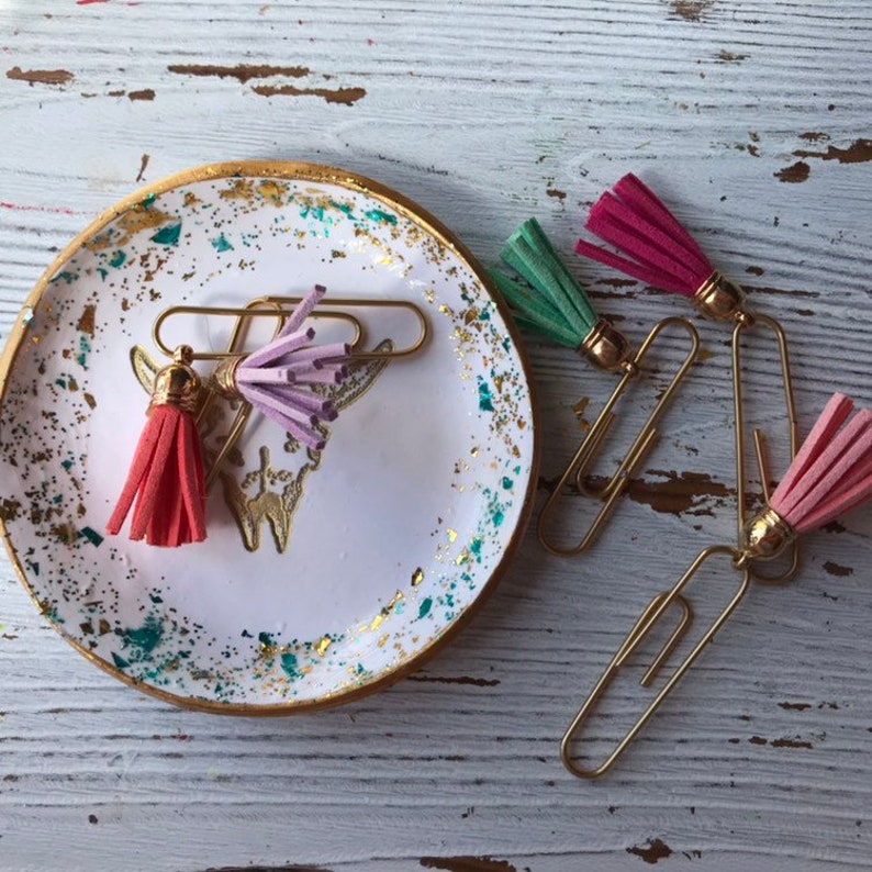 May include: Four gold paper clips with colorful tassels attached. The tassels are pink, green, and purple. The paper clips are sitting on a white surface with gold flecks.