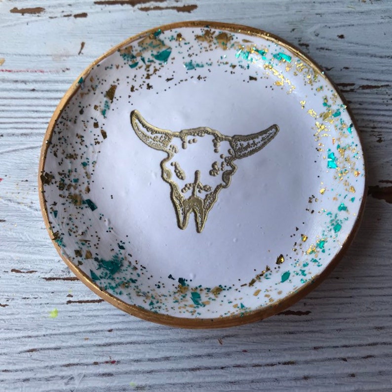 May include: White ceramic trinket dish with a gold rim and a gold glitter design. The dish features a white embossed bull skull design in the center.