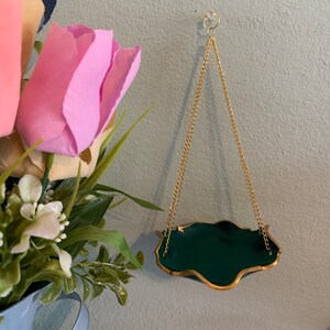 Op de afbeelding: Een donkergroene hangende plantenpot met een gouden rand en een gouden ketting. De pot hangt aan een haak aan een lichtblauwe muur.