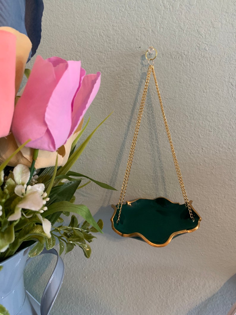 Op de afbeelding: Een donkergroene hangende plantenpot met een gouden rand en een gouden ketting. De pot hangt aan een haak aan een lichtblauwe muur.