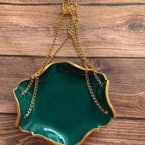 Op de afbeelding: Een groene keramische hangende plantenpot met een gouden rand en een gouden ketting. De pot heeft een golvende rand en hangt aan een gouden ring.