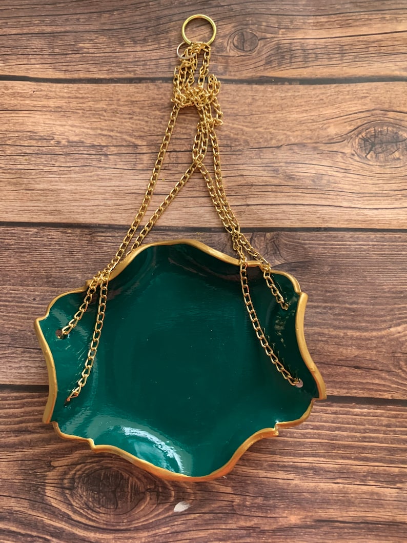 Op de afbeelding: Een groene keramische hangende plantenpot met een gouden rand en een gouden ketting. De pot heeft een golvende rand en hangt aan een gouden ring.