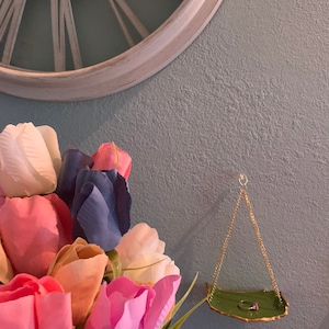 K&ouml;nnte beinhalten: Eine gr&uuml;ne Keramik-Ringhalterung mit goldenem Rand h&auml;ngt an einer goldenen Kette. Die Schale ist blattf&ouml;rmig und hat einen Ring darauf liegen. Die Schale h&auml;ngt an einer hellblauen Wand mit einem Strau&szlig; rosa, wei&szlig;er und blauer Tulpen im Vordergrund.