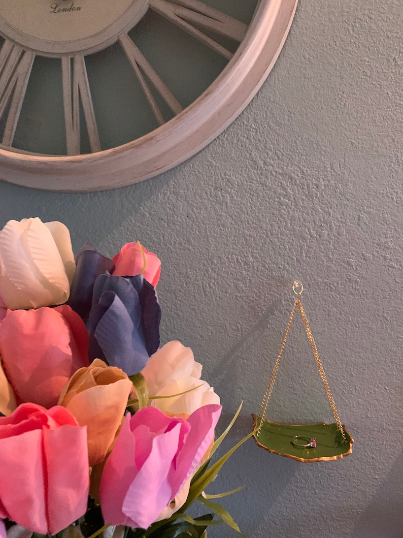 K&ouml;nnte beinhalten: Eine gr&uuml;ne Keramik-Ringhalterung mit goldenem Rand h&auml;ngt an einer goldenen Kette. Die Schale ist blattf&ouml;rmig und hat einen Ring darauf liegen. Die Schale h&auml;ngt an einer hellblauen Wand mit einem Strau&szlig; rosa, wei&szlig;er und blauer Tulpen im Vordergrund.