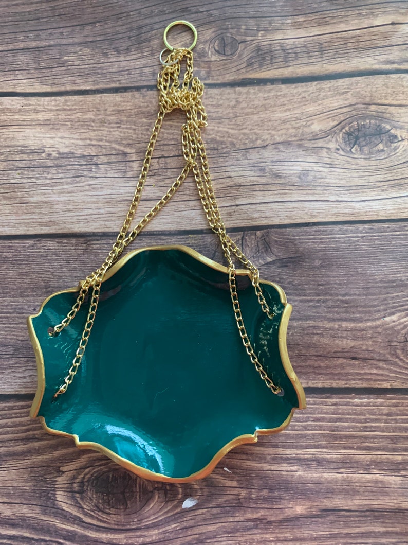 Op de afbeelding: Een donkergroene hangende schaal met een gouden rand en een gouden ketting. De schaal hangt aan een gouden ring.