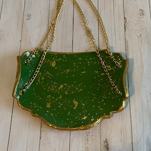 K&ouml;nnte beinhalten: Ein gr&uuml;nes und goldenes H&auml;ngeschild mit einer goldenen Kette. Das Schild hat eine gesprenkelte gr&uuml;ne Oberfl&auml;che mit goldenen Glitzerakzenten.