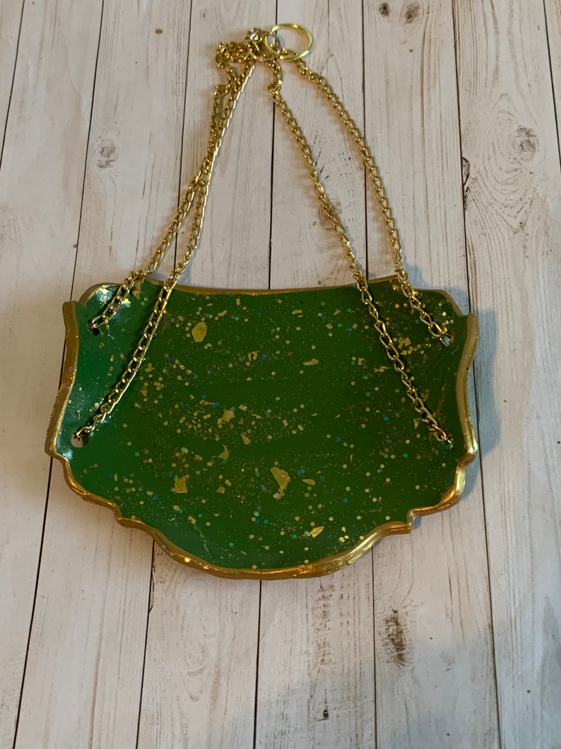 K&ouml;nnte beinhalten: Ein gr&uuml;nes und goldenes H&auml;ngeschild mit einer goldenen Kette. Das Schild hat eine gesprenkelte gr&uuml;ne Oberfl&auml;che mit goldenen Glitzerakzenten.