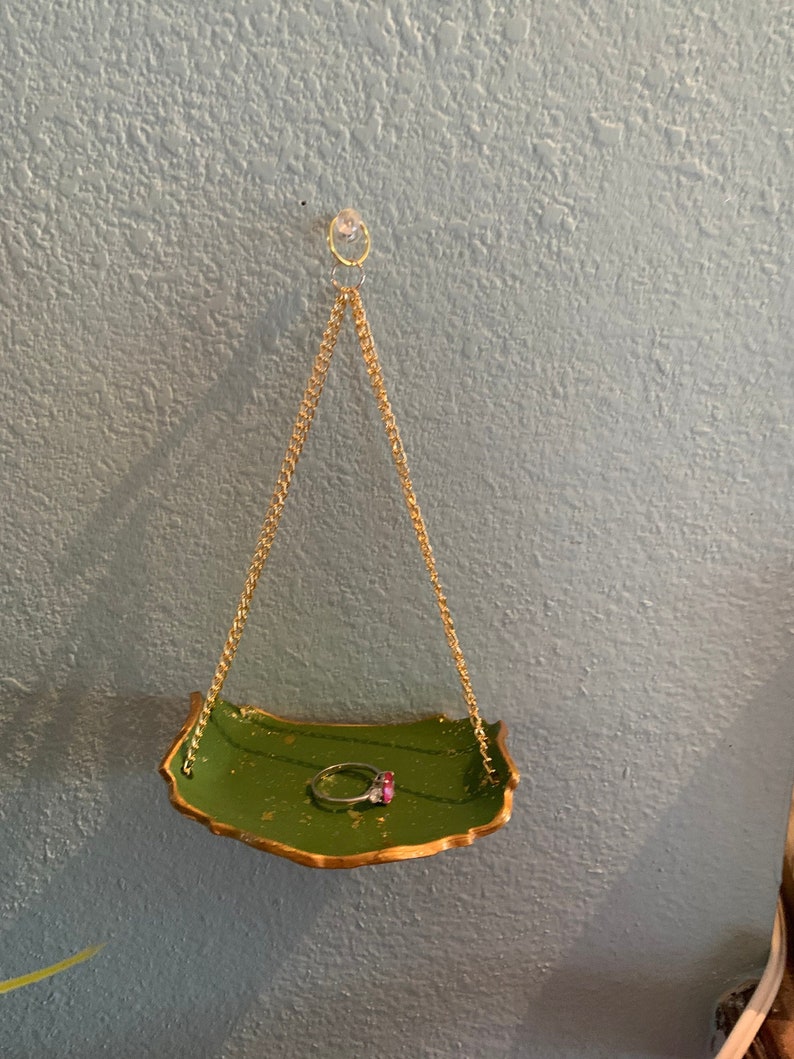 K&ouml;nnte beinhalten: Ein gr&uuml;ner Ringhalter mit goldenen Akzenten h&auml;ngt an einer goldenen Kette. Der Halter ist an einem Haken an einer hellblauen Wand befestigt. Ein Ring mit einem roten Stein liegt im Halter.