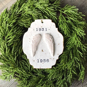May include: White ceramic ornament with gold flecks, featuring a pair of angel wings and the dates 1931-1956.