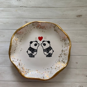 May include: White ceramic ring dish with a gold rim and a glittery finish. Two black and white panda bears are kissing with a red heart between them. The initials "S" and "M" are above the bears.