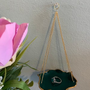 Op de afbeelding: Een groene en gouden ringhouder hangt aan een gouden ketting. De schaal heeft een gegolfde rand en hangt aan het plafond. Een ring met een grote smaragdgroene steen ligt in de schaal.