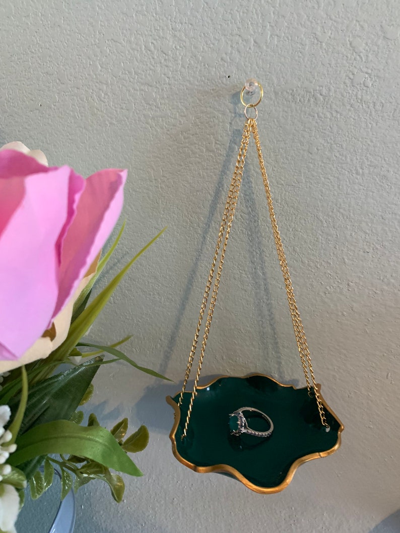 Op de afbeelding: Een groene en gouden ringhouder hangt aan een gouden ketting. De schaal heeft een gegolfde rand en hangt aan het plafond. Een ring met een grote smaragdgroene steen ligt in de schaal.