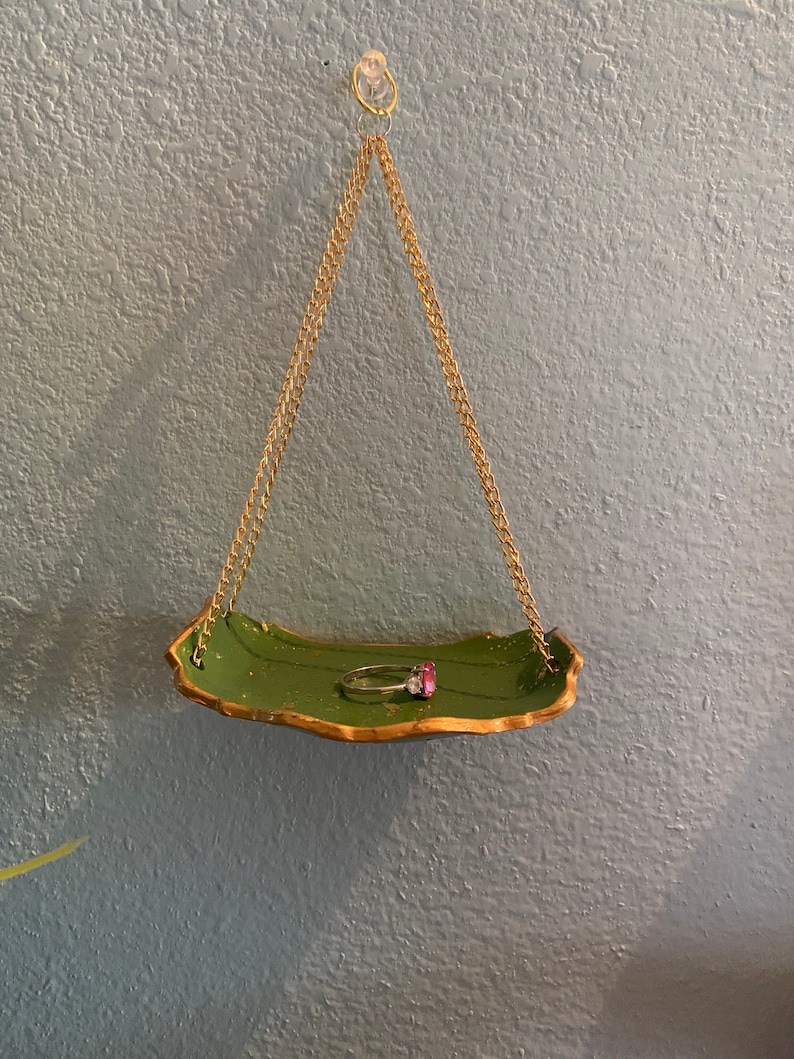K&ouml;nnte beinhalten: Ein gr&uuml;nes und goldenes, blattf&ouml;rmiges Tablett h&auml;ngt an einer goldenen Kette. Auf dem Tablett liegt ein Ring mit einem rosafarbenen Stein.
