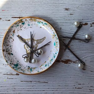 May include: A white ceramic dish with a gold rim and a skull design. The dish is filled with hair clips with pearl accents. The dish is sitting on a white wooden surface.