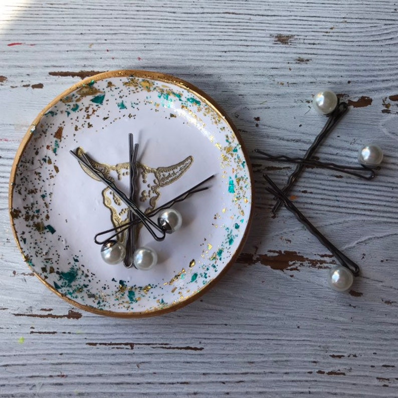 May include: A white ceramic dish with a gold rim and a skull design. The dish is filled with hair clips with pearl accents. The dish is sitting on a white wooden surface.